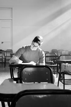 Monochrome photo of a young man studying in an empty classroom with natural light streaming in from a window.