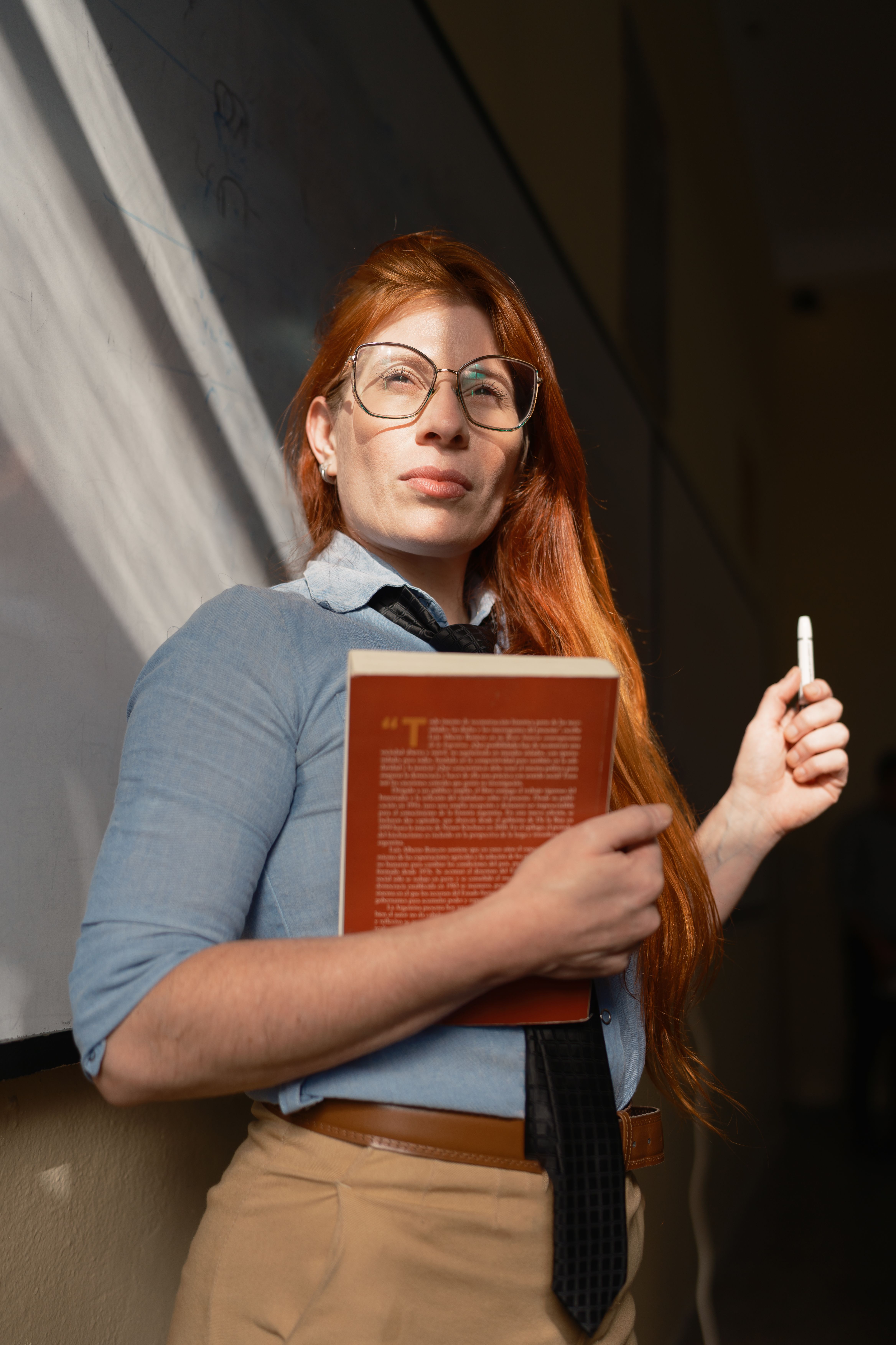 A red-haired instructor confidently holds a book while standing by a classroom board.
