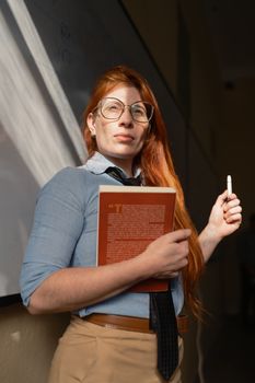 A red-haired instructor confidently holds a book while standing by a classroom board.