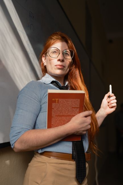 A red-haired instructor confidently holds a book while standing by a classroom board.
