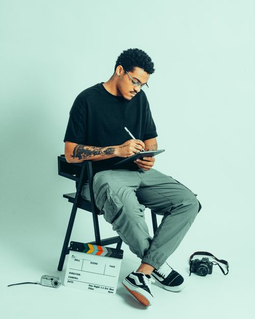 Free Filmmaker seated with tablet, surrounded by camera gear and clapperboard, signifying creative process. Stock Photo