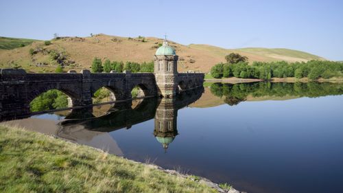 Free A scenic view of a historic stone bridge reflecting in a calm lake under a clear sky. Stock Photo