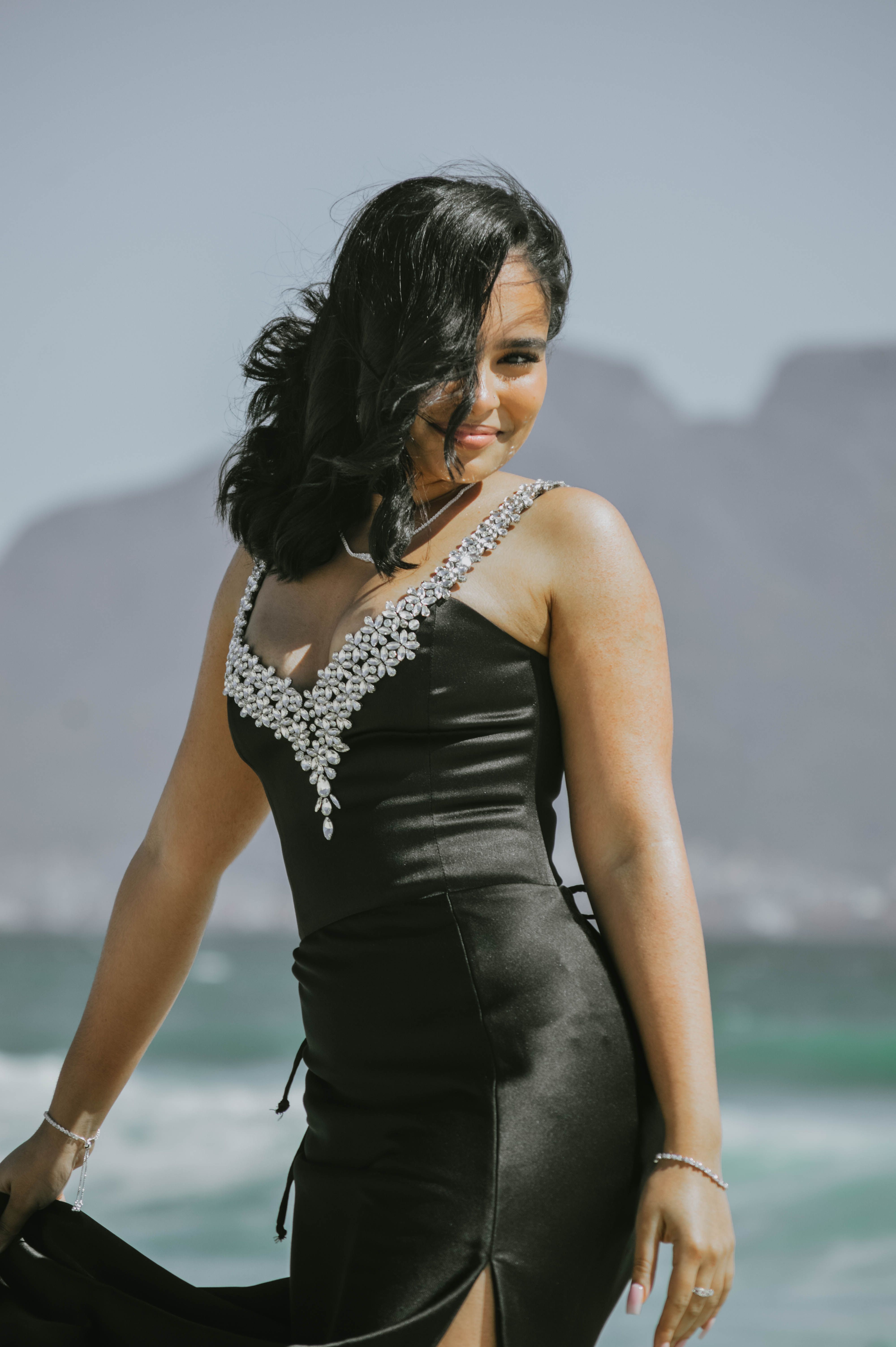 A beautiful woman in a black dress adorned with jewels stands by the ocean, with mountains in the background.