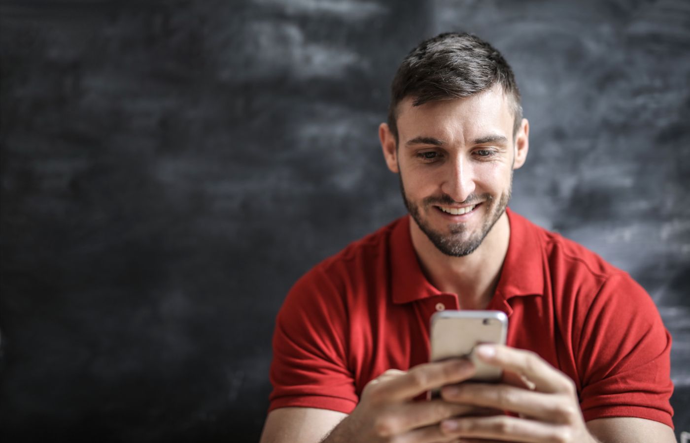 A man in a red polo shirt happily using his smartphone indoors against a dark background.