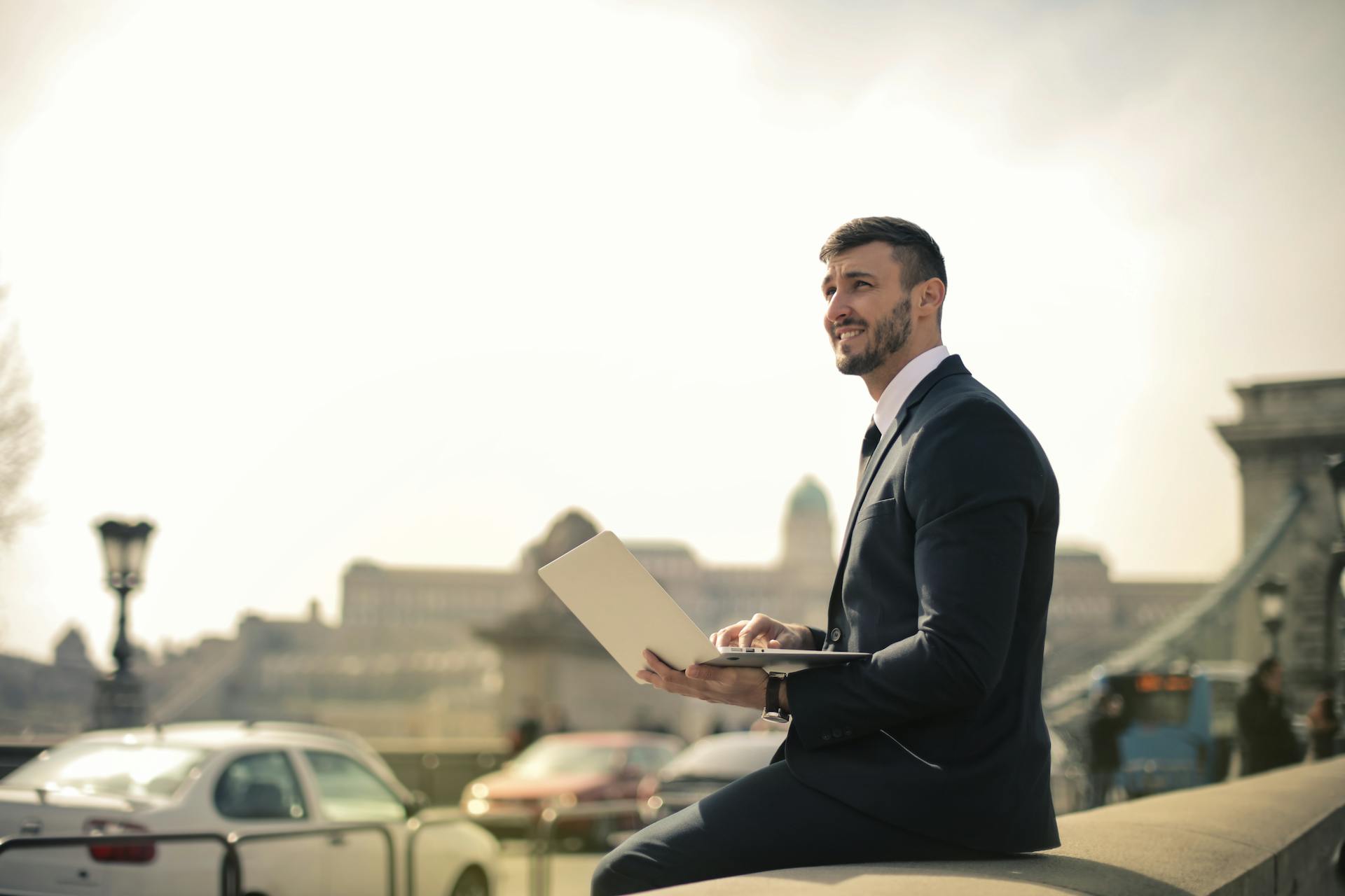 Businessman in formal attire sitting outdoors with a laptop, smiling in a city setting.