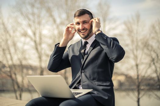 Smiling businessman in formal attire using headphones and laptop outdoors.