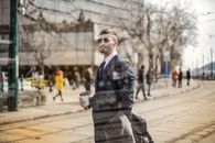 Through glass view of bearded man in suit and eyeglasses looking away while standing with coffee on city street