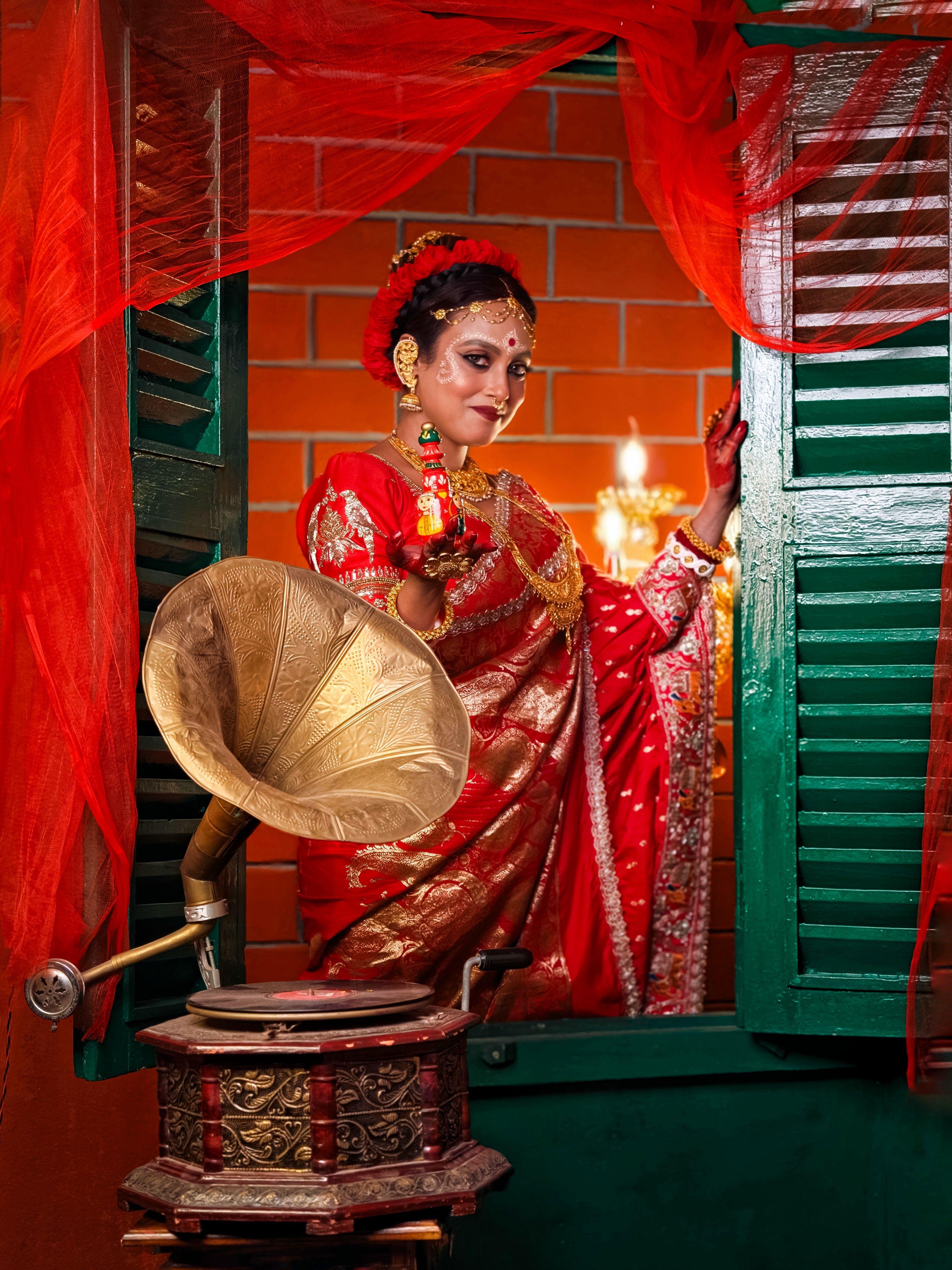 A stunning Indian bride in red sari poses beside a vintage gramophone near a window.