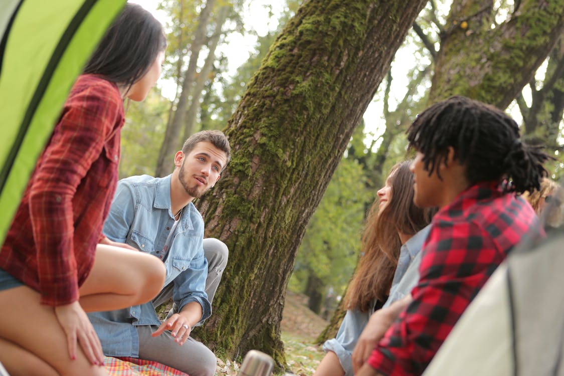 Free Group of young people in casual clothes sitting on plaid around near trees with moss while talking and looking at each other Stock Photo