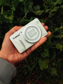 A hand holds a compact Canon camera amidst lush green foliage outdoors.