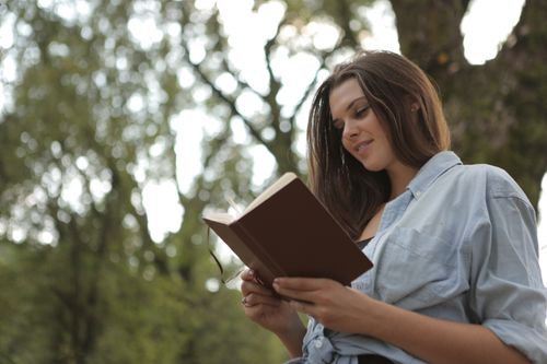 Free Young woman reading a book outdoors, surrounded by nature, looking content. Stock Photo