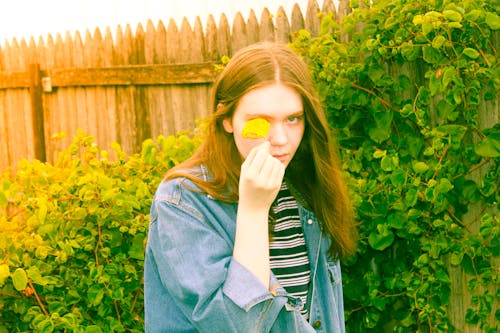 Free Young casual female teenager in denim shirt covering eye with yellow flower looking at camera in summer garden Stock Photo