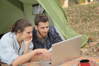 Cheerful female and bearded male using laptop with touchpad while resting in tent at campsite