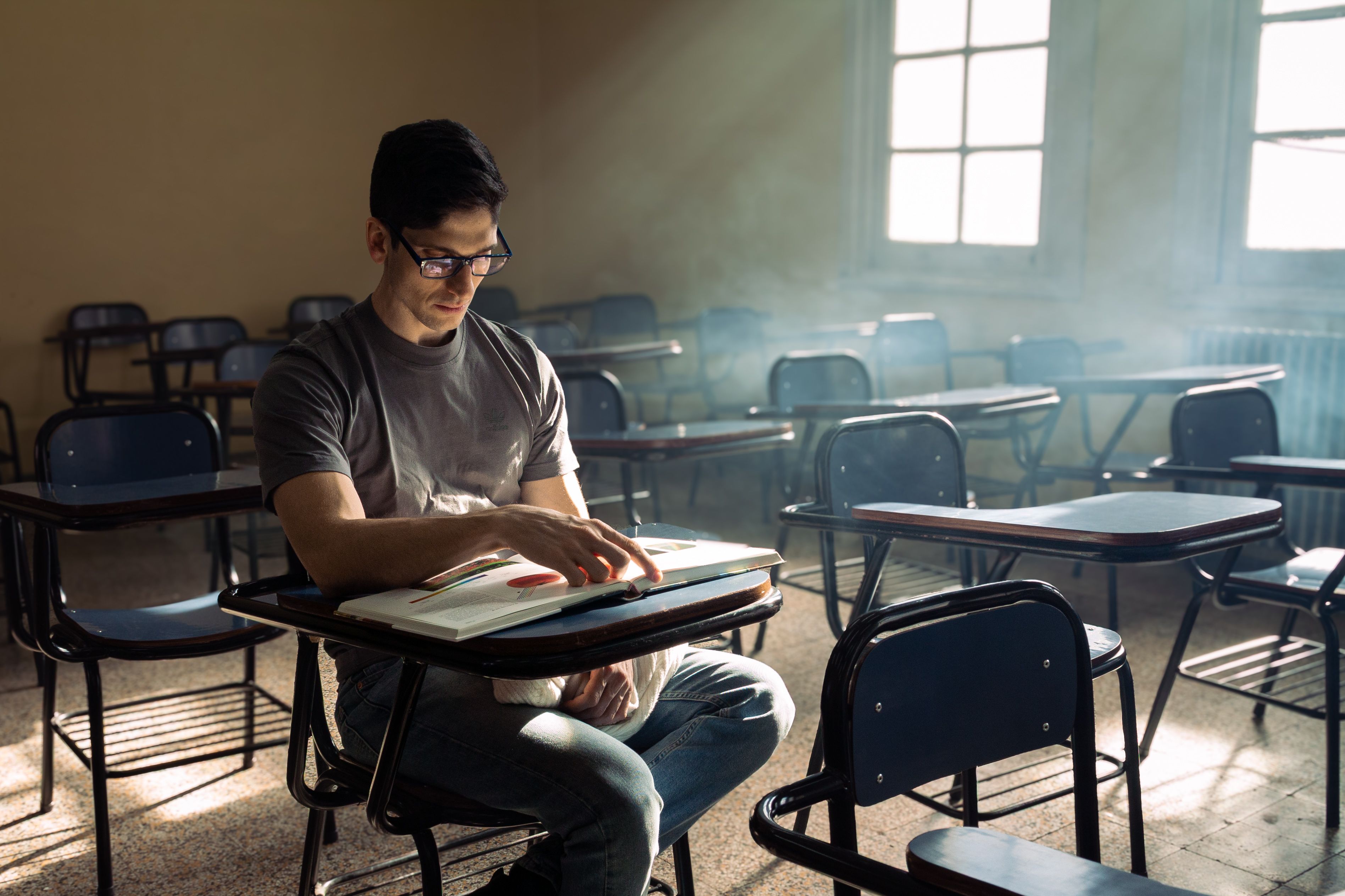 A young man reading alone in a sunlit classroom, surrounded by empty desks.