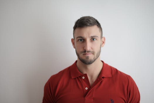 一名神情严肃、穿着红色马球衫的男子倚着白墙摆姿势