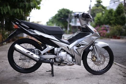 Silver motorbike parked outdoors on a quiet street, surrounded by greenery and trees.