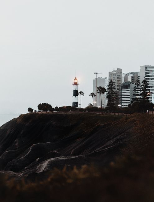 A lighthouse stands on a rocky cliff in Lima, Peru, against a backdrop of modern buildings, capturing the essence of coastal serenity.