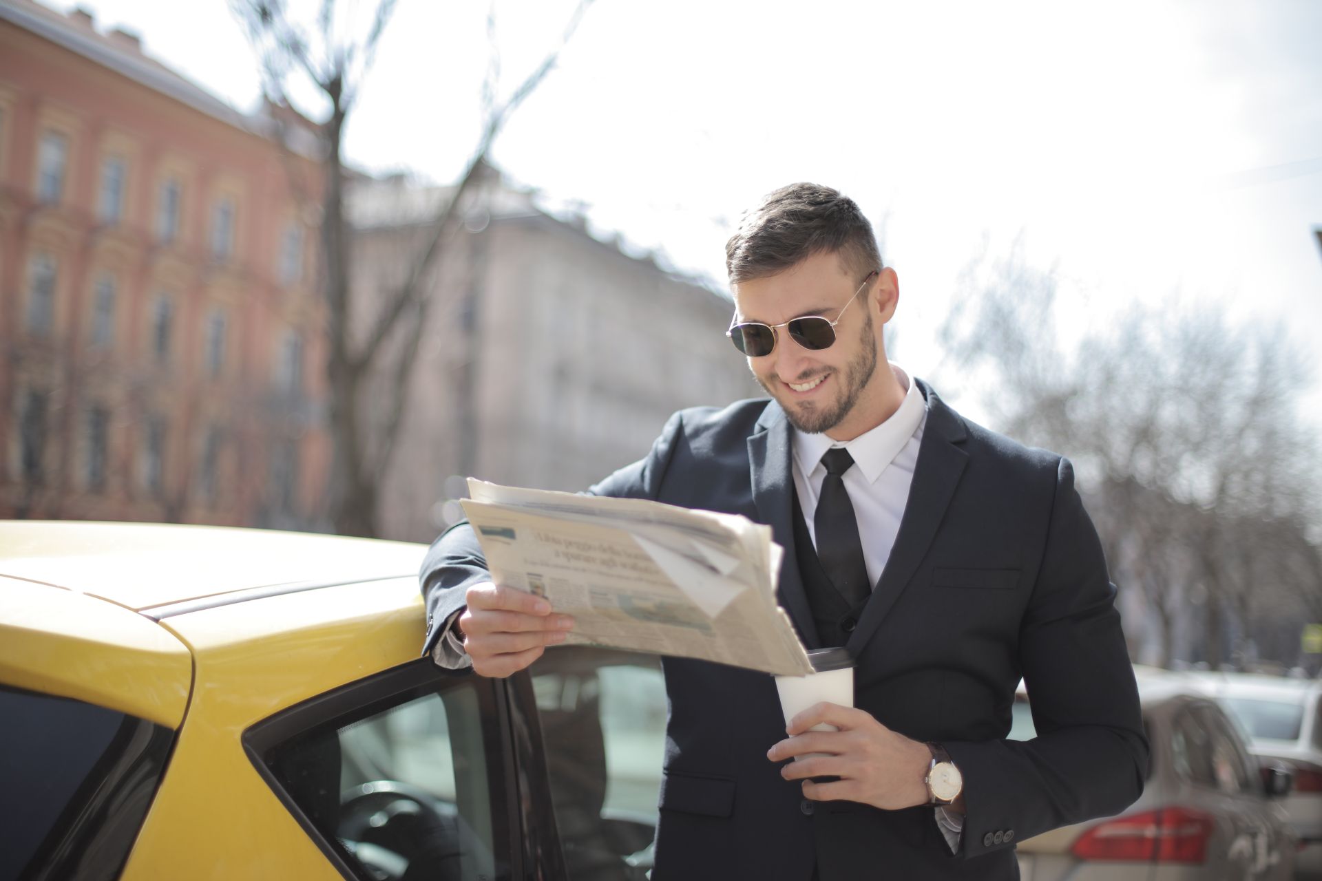 A stylish businessman in a suit reads a newspaper outside by a car, holding a coffee cup.