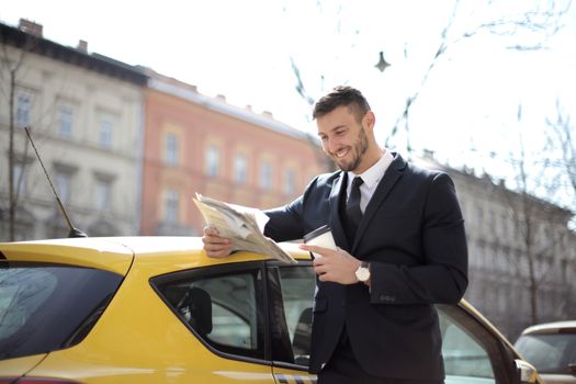 Smiling businessman in a suit reading a newspaper and having coffee by a car on a sunny day.