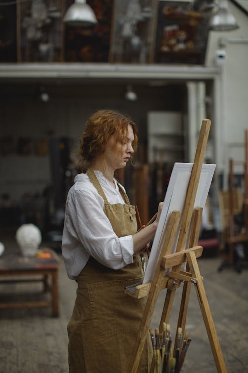Женщина в белой рубашке с длинным рукавом и коричневом фартуке стоит во время рисования