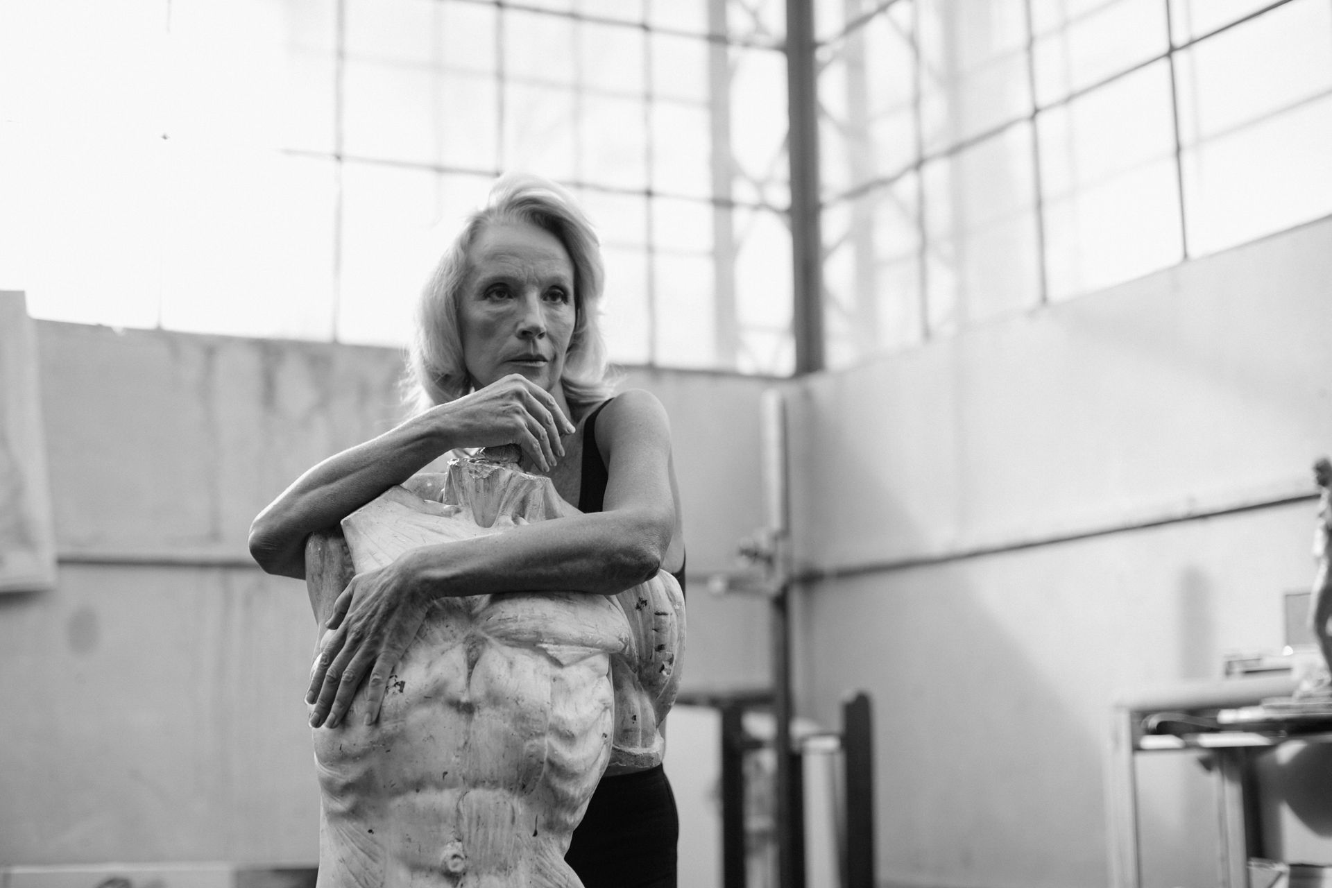An elderly woman in deep thought posing with a torso sculpture in an artist's studio.