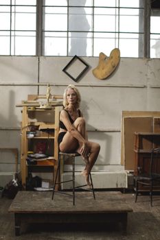 A woman in lingerie posed elegantly on a stool in an art studio, creating a classic and contemplative atmosphere.
