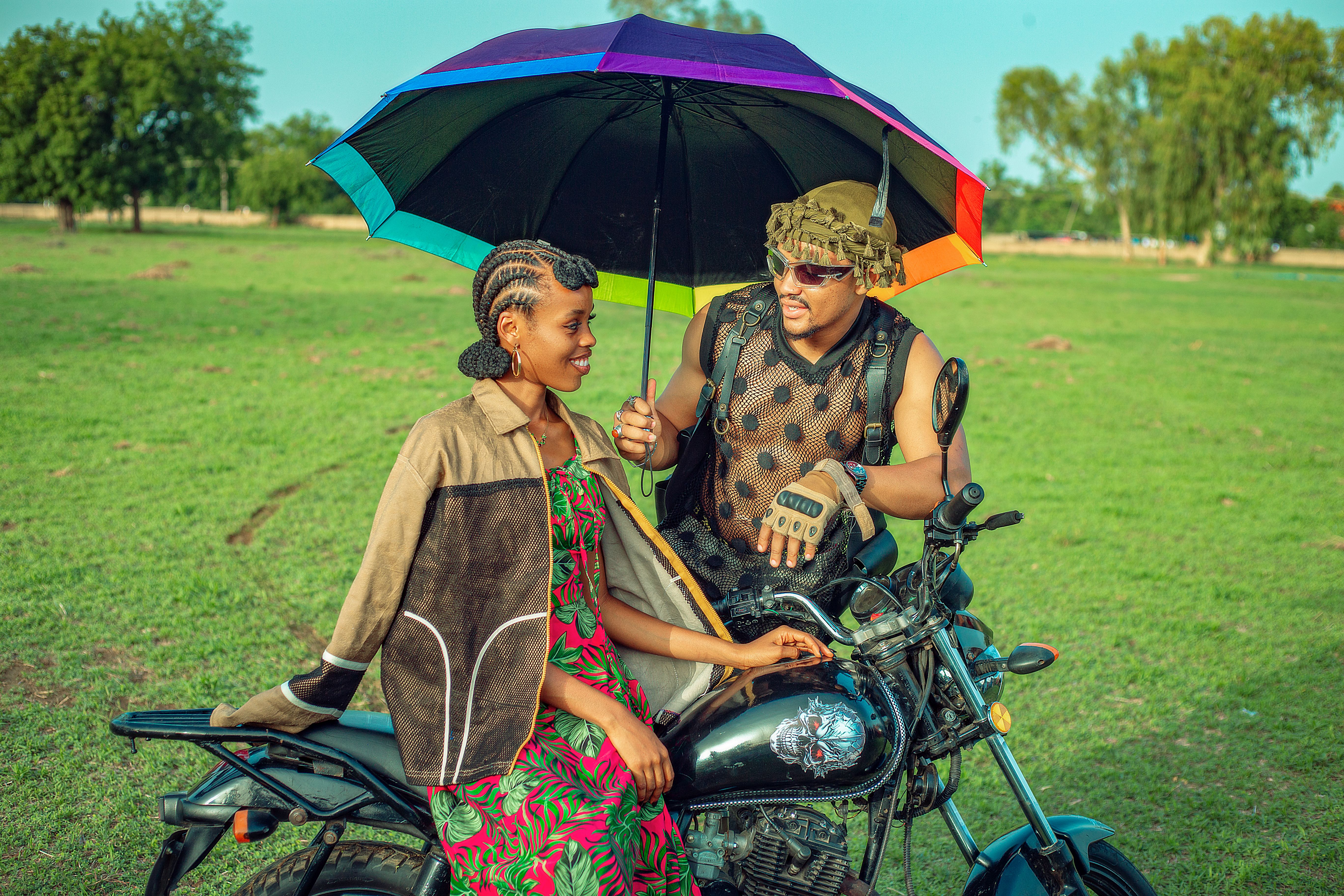 A stylish couple on a motorcycle under a colorful umbrella, enjoying a sunny day outdoors.