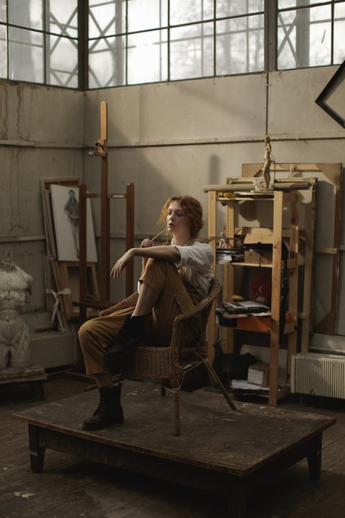 Free A contemplative woman sitting on a chair in an artist's studio surrounded by sculptures and art materials. Stock Photo