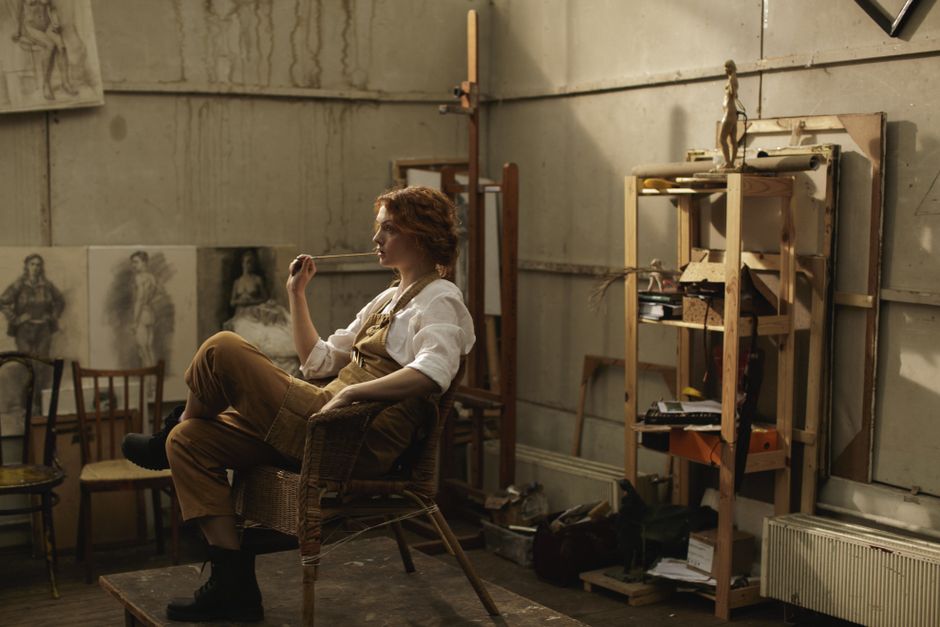 Female artist in a lofty art studio, lost in thought with a paintbrush, surrounded by drawings.