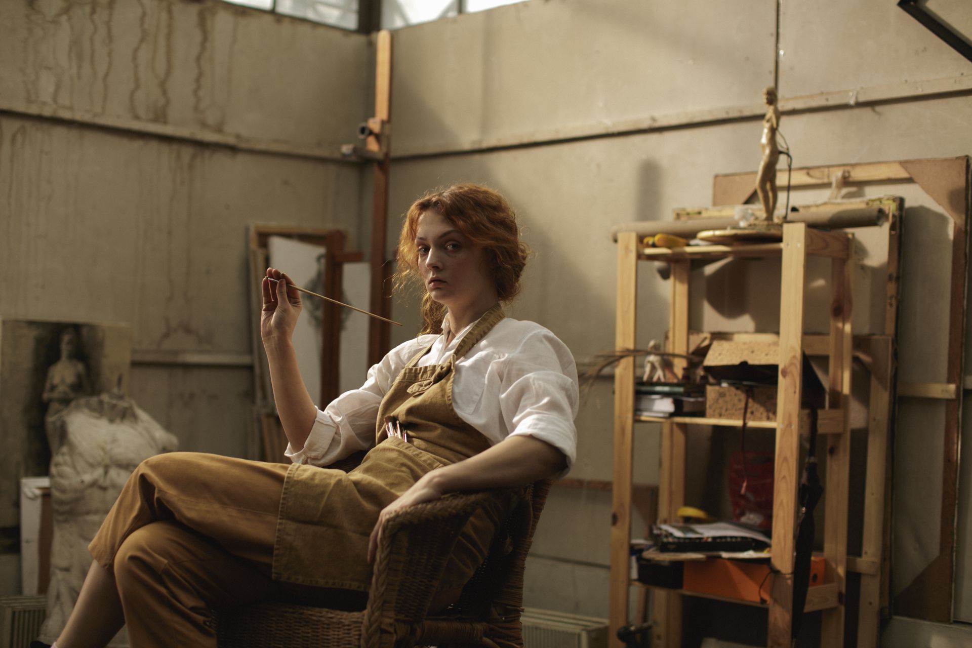 A woman artist seated in a workshop, surrounded by art materials and sculptures, conveying creative concentration.