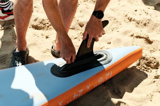 Close-up of man fitting a paddleboard fin on a sunny beach. Adventure and watersport concept.