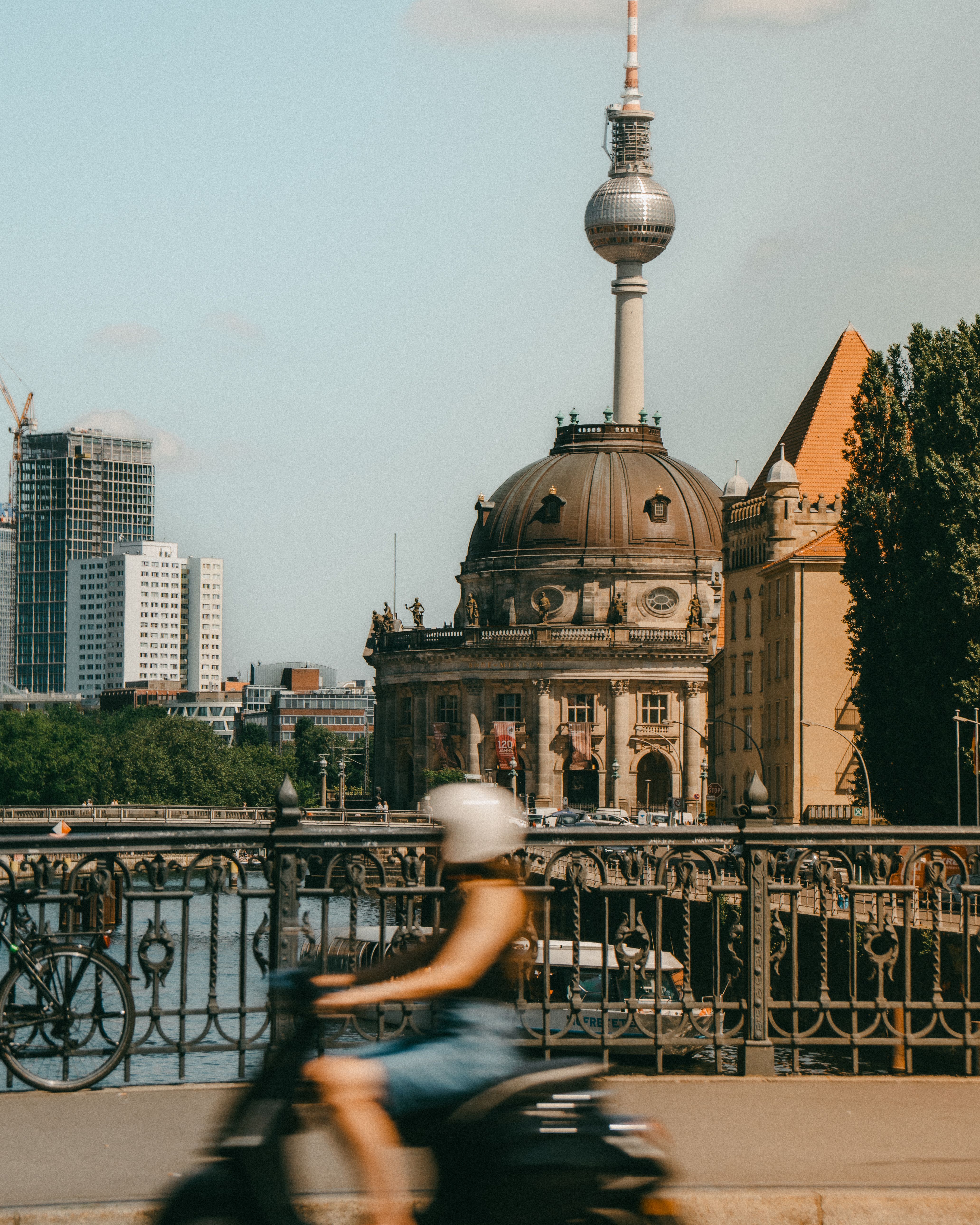 A woman on a scooter rides across a bridge in front of the iconic Berlin TV Tower and Bode Museum.