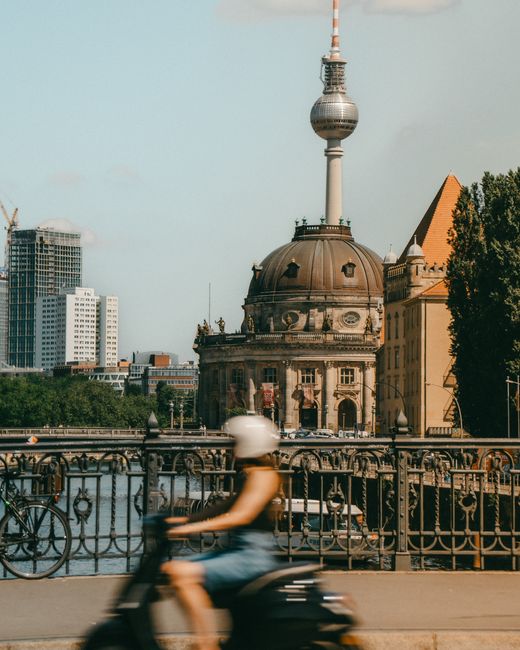 A woman on a scooter rides across a bridge in front of the iconic Berlin TV Tower and Bode Museum.