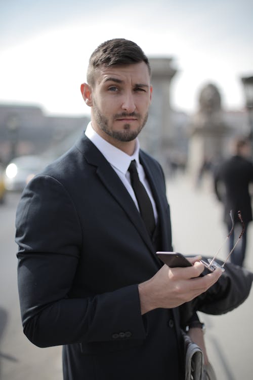 Man in Black Suit Holding Smartphone and Sunglasses