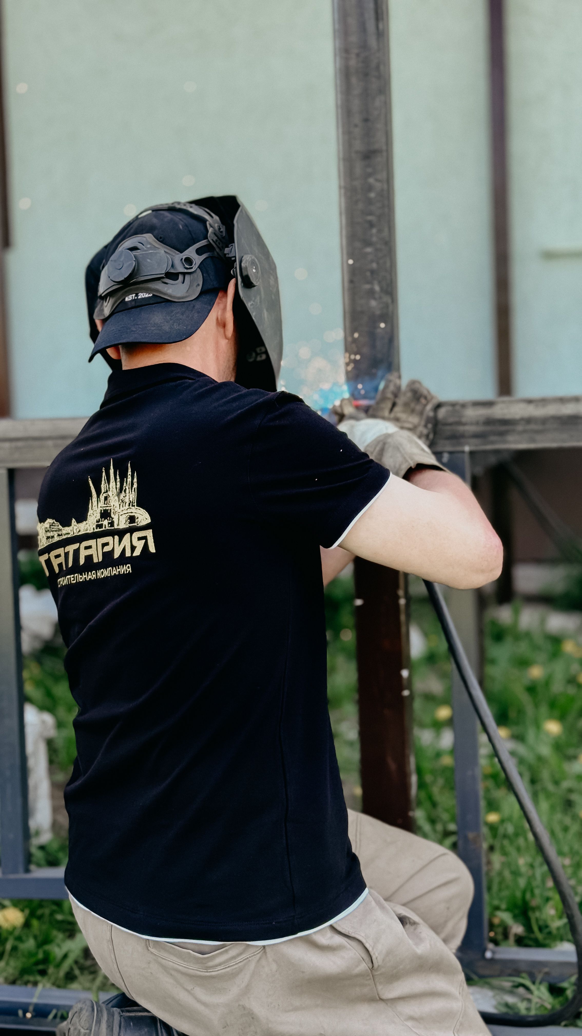 A welder wearing a helmet and gloves works outdoors on metal construction.