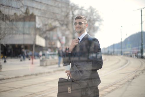 Free A confident businessman walks in the city, holding a coffee cup and a briefcase, wearing sunglasses. Stock Photo