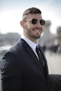 一位穿着黑色西装、戴着太阳镜的商人，在户外自信地微笑着，现代发型更显自信