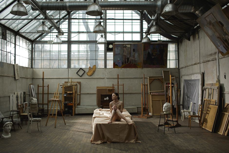 A woman poses nude in an industrial art studio, surrounded by creative tools and sculptures.
