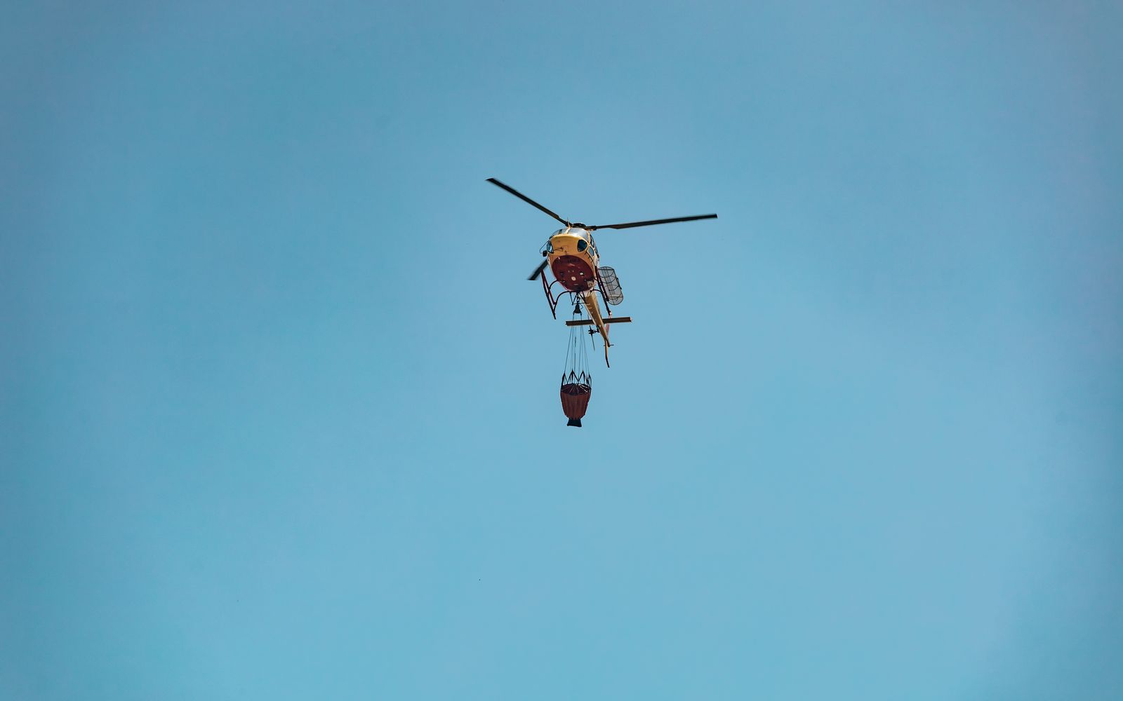 A firefighting helicopter flying with a water bucket suspended beneath it against a clear sky