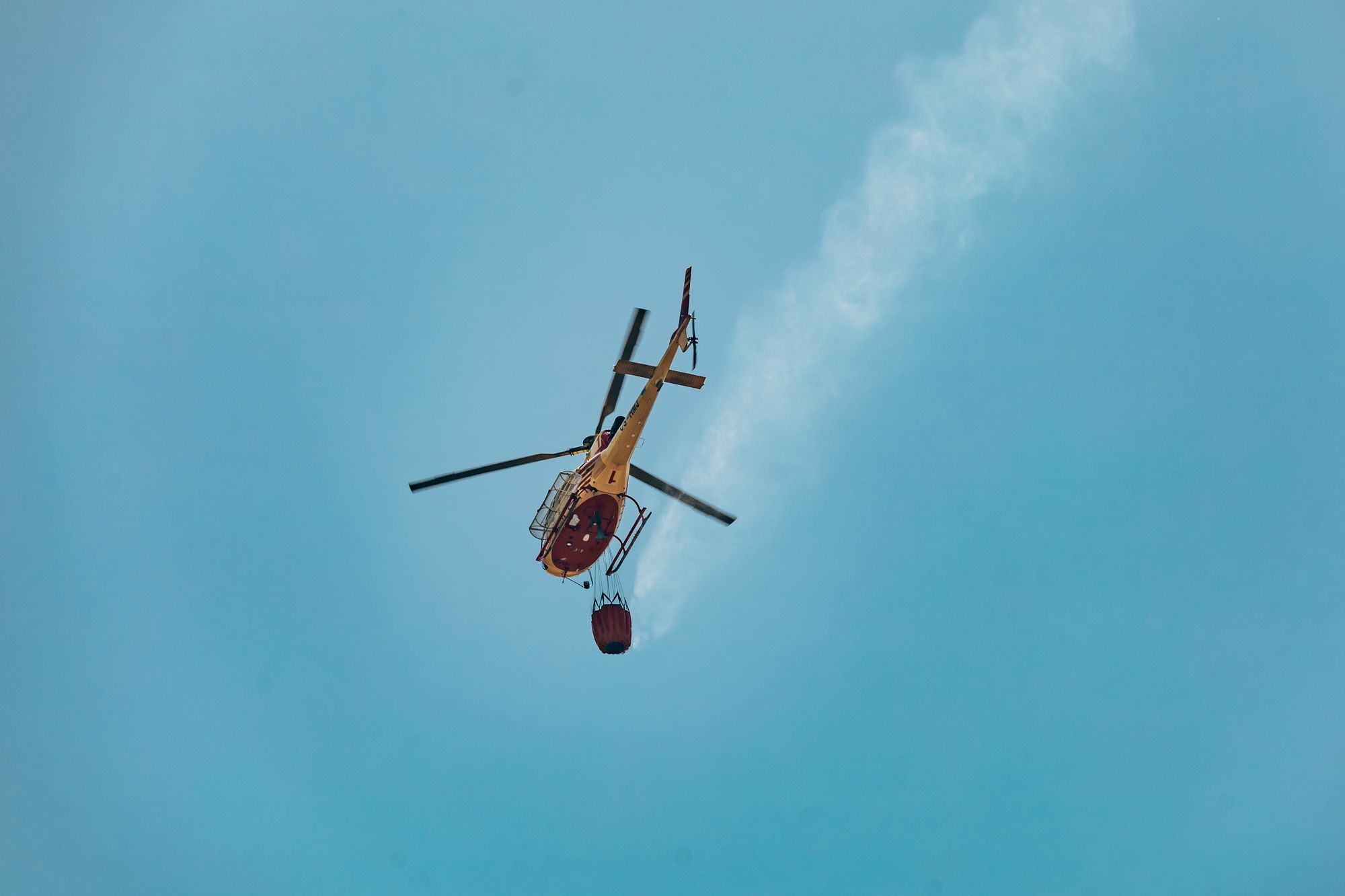 Helicopter performing aerial firefighting with water bucket against a clear sky in Braga, Portugal.