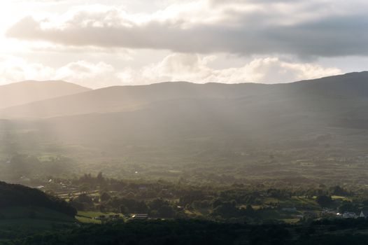 A serene landscape view of Kilgarvan, Ireland, with sunlight breaking through clouds over rolling hills.