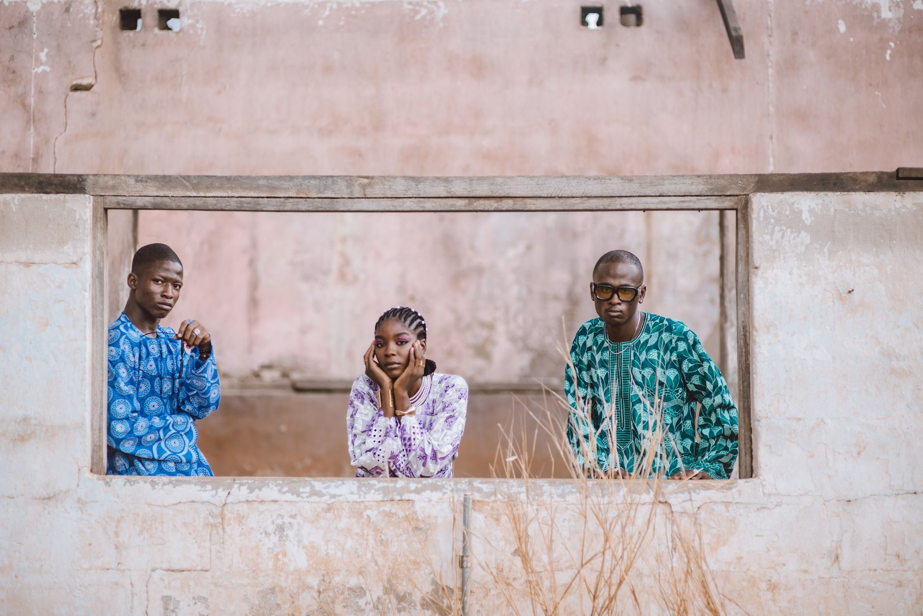 Three individuals pose in colorful African attire within a rustic frame outdoors.