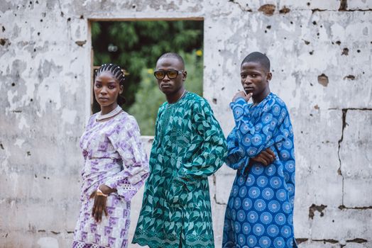 Three adults in colorful African attire pose confidently against a rustic wall, showcasing vibrant cultural fashion.