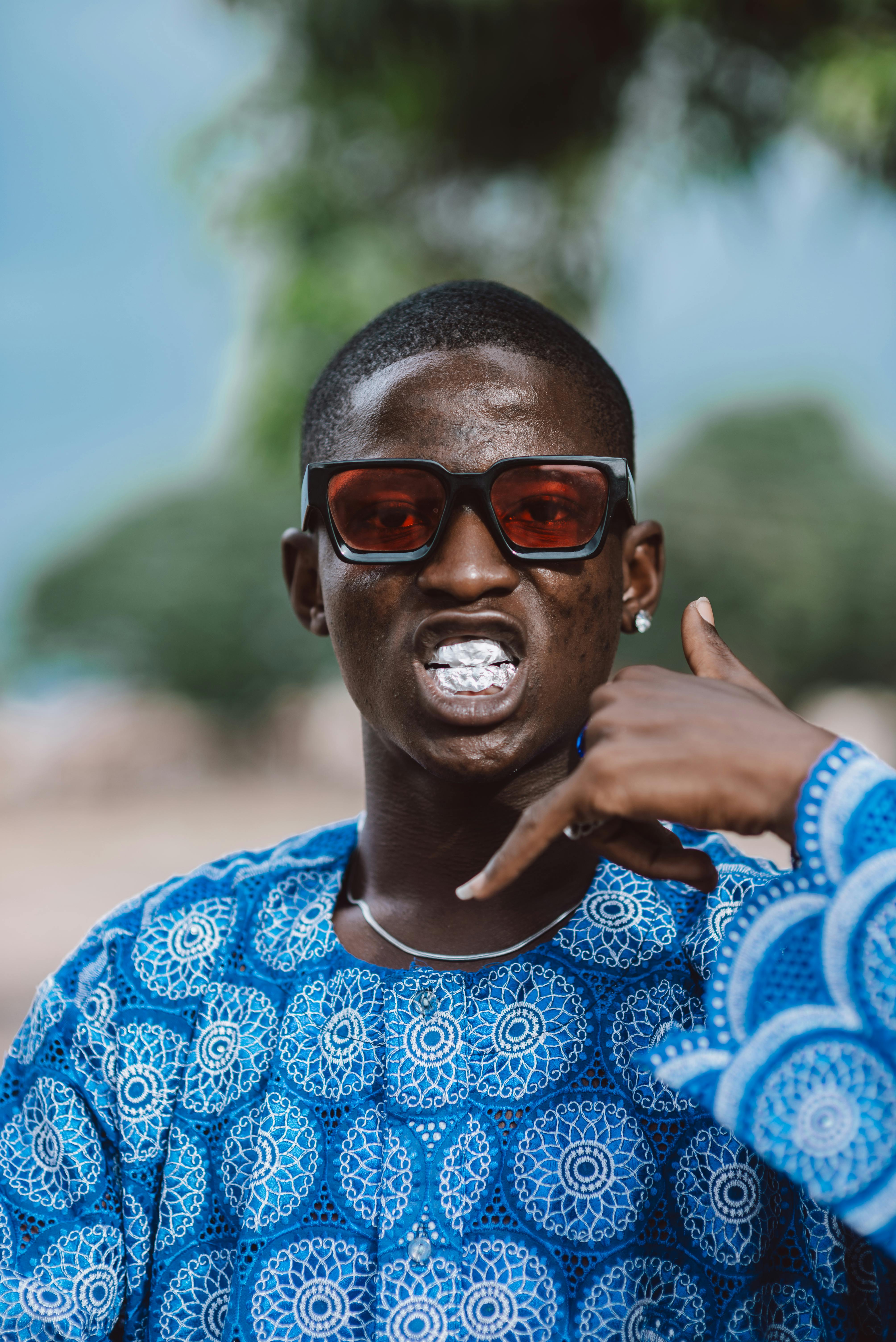 A fashionable individual wearing blue traditional attire and sunglasses making a phone gesture outdoors.