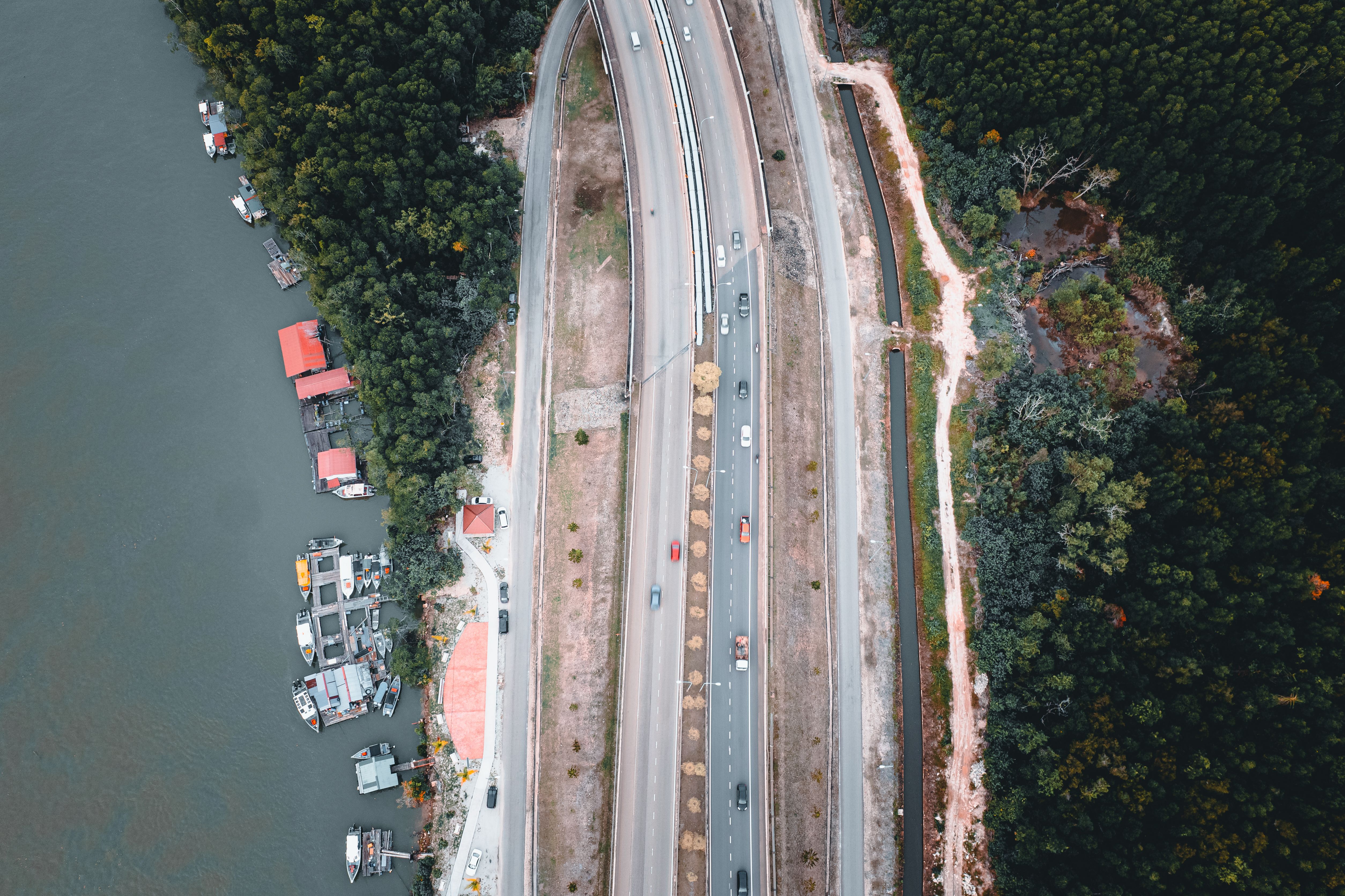 Aerial view of a highway running alongside a river and lush forest, with boats docked nearby.
