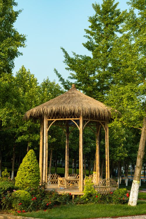 Free A charming bamboo gazebo with a thatched roof surrounded by lush greenery in a sunlit park. Stock Photo