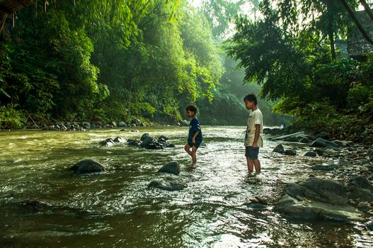 Two children explore a serene river surrounded by lush greenery in Banten, Indonesia.