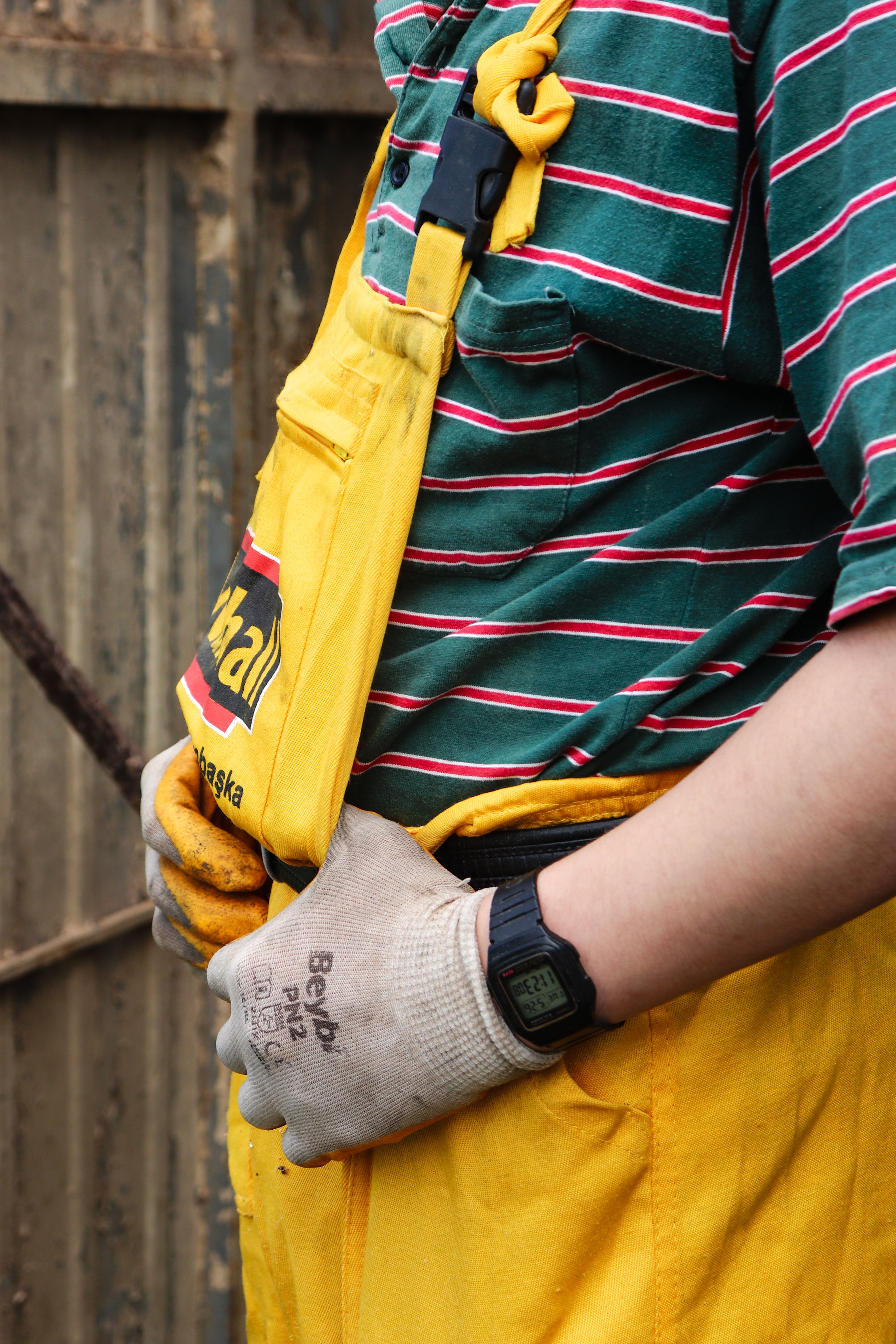 Close-up of a worker wearing a striped shirt and yellow overalls with a glove and watch, outdoors.