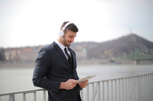 Man in Black Suit Jacket and Black Pants Holding an Tablet While Standing Beside Railings