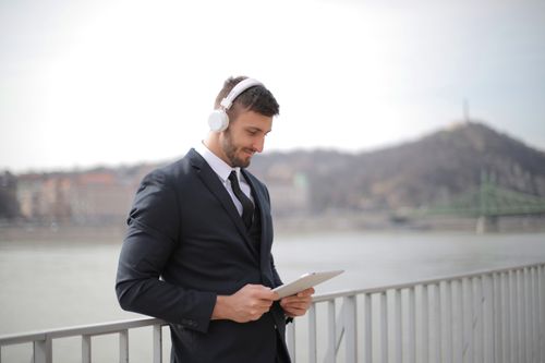 Free A businessman in a suit using a tablet while listening to music near a river in an urban setting. Stock Photo